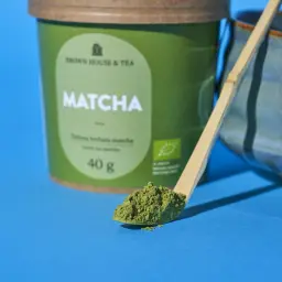 Zielone matcha na łyżeczce, idealne jako herbata na odchudzanie. Obok zielony pojemnik z napisem "Matcha" i niebieska miseczka.