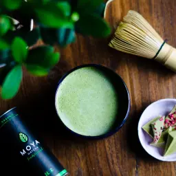 Szukasz, gdzie kupić matcha stacjonarnie? Na zdjęciu widać puszkę Moya Matcha, miseczkę z zieloną czekoladą i bambusową miotełkę.